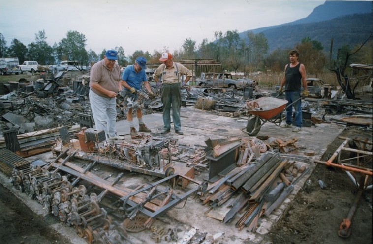 Three men sort through twisted metal. A fourth wheels a wheel barrow.