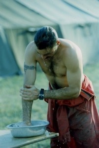 Man stripped to the waist washing his arm in a basin on a table. Hair is wet. Coveralls are dirty. Tents in background