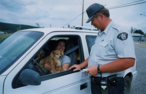 RCMP officer with printed information talking to a woman in a white vehicle at the side of a road. Driver has a dog in her lap.