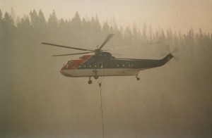 Helicopter in flight in a smoke filled sky. Cable hangs from the aircraft. Forest in background.