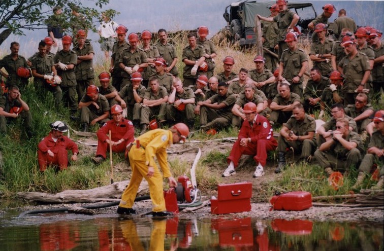 Group of soldiers and forestry personnel watch a man in yellow coveralls operate a water pump. Most wear hard hats. Water in foreground.