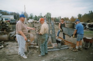 Group of men sort through metal. One holds a wheel. Two others carry a large metal box. Twisted metal in foreground.