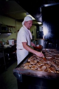 Cook in white chef’s hat, kerchief, and clothing grilling pork chops on a grill.