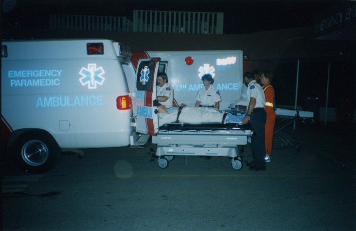 Three ambulance attendants loading a person on a stretcher into an ambulance. Emergency worker in orange outfit with reflective stripes looks on.