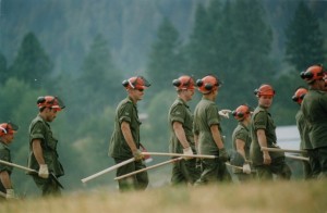 Group of men in green army uniforms walking through a field wearing orange hard hats and carrying shovels and grub hoes. One soldier looks at the camera.