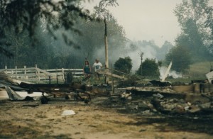 Couple walks through twisted metal, rubble and a foundation. Smoke in background.