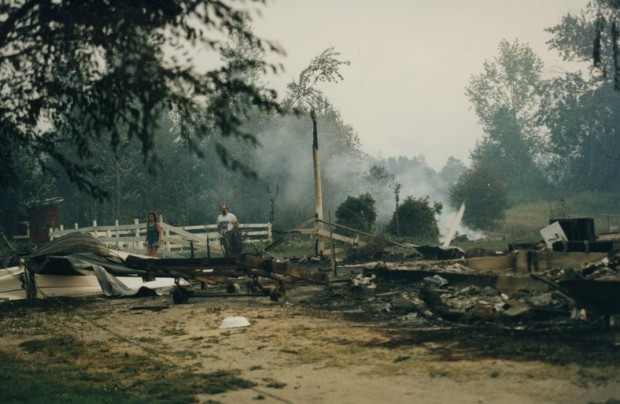 Couple walks through twisted metal, rubble and a foundation. Smoke in background.