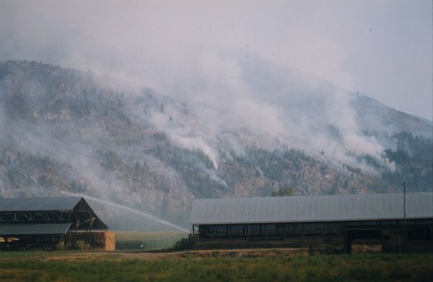Fire burning in the hills behind a farm. Hay barns in foreground. Water being sprayed on fields.