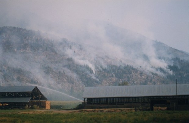 Fire burning in the hills behind a farm. Hay barns in foreground. Water being sprayed on fields.