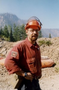 Man with a goatee and glasses wearing a hard hat. Forest and hills behind him.