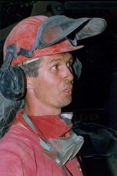 Man in a red shirt with a mask and goggles around his neck, wearing a hard hat talking.