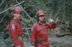 Two men in red coveralls and orange hard hats, with a third man hidden behind them, look off into the distance. Man with mustache motions to someone out of the picture. Forest in background.