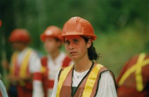 Young woman with dark hair, wearing a safety vest and hard hat stares into distance.