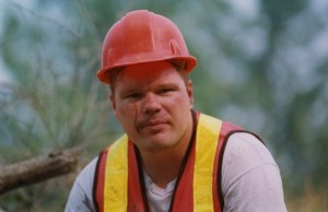Man in a safety vest and a hard hat with dirt streaking his face looks at the camera.