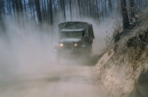Covered army truck travels on dusty road in a burned forest.