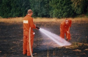 Deux hommes en combinaison orange. L'un d'eux tient un boyau d'arrosage et asperge le sol pendant qu'un autre creuse.