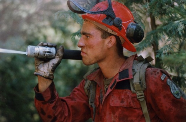 Man in coveralls and a helmet holds a hose over his shoulder and sprays something out of sight.