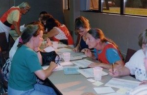 Woman in a red pinny and braided hair leans forward towards a woman wearing a blue/green shirt. Eight other people are busy with paperwork.