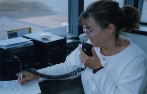 Assise à un bureau, une femme coiffée d'une queue de cheval et portant des lunettes inscrit des informations sur une feuille et parle dans un micro.