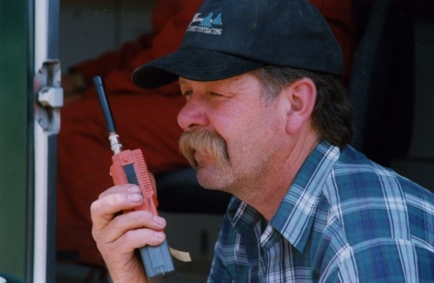 Man in a blue and white checked shirt and blue baseball cap speaking into a hand held radio.