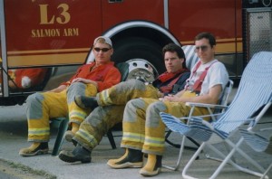 Three men in firefighter’s pants and rubber boots sit in lawn chairs beside a fire engine labeled “L3”.