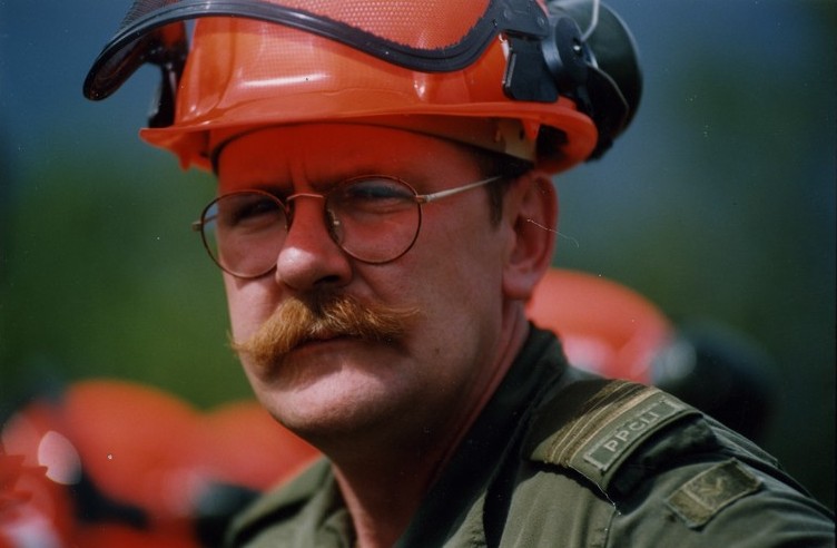 Mustached man in green uniform, wearing a hard hat and glasses squints at the camera.