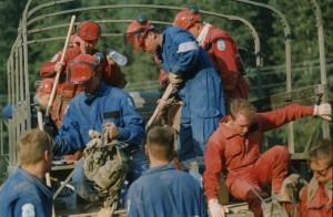 Men in coveralls, some with hard hats, carrying shovels and backpacks, getting out of an uncovered army truck.