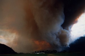 Smoke from fire billows from hills. Sky is lit with orange. Farm in centre of photo.