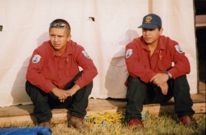 First Nations firefighters dressed in red shirts and black pants sit on wooden tent platform as sun sets.