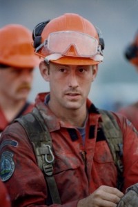 Man wearing dirty coveralls and a hard hat looks away from camera.