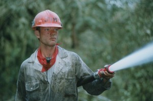 Man wearing dirty coveralls and a hard hat spraying water through hose.