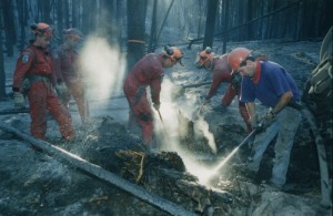 Two firefighters in red coveralls dig in the burned ground. A man with a purple t-shirt sprays water into the ground. Two men in red coveralls look on. All men wear hard hats.