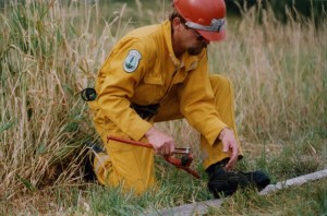 Man in yellow coveralls, red hard hat, tool in hand, foot on hose.