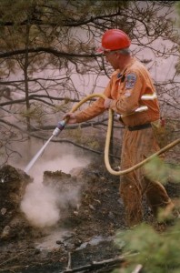 Man in yellow coveralls and hard hat sprays hose into fire.