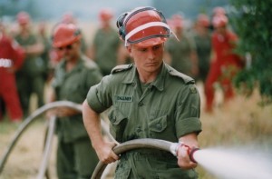 Soldier in green uniform, wearing hard hat, holds hose spraying water.