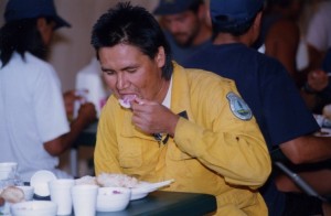 Man in yellow overalls sits at a table eating a meal.