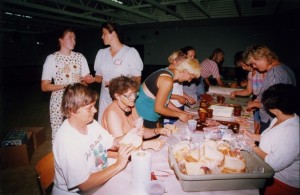Group of women standing and sitting around a table, making sandwiches. Two women stand in background.