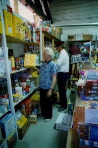 Older woman puts box of food on a shelf at food bank. Two other people also stock shelves in the background.