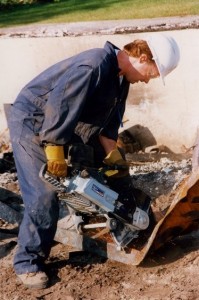 Man wearing a hard hat and blue coveralls operates a metal saw. Rubble in background.