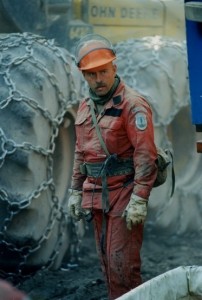 Man wearing red coveralls and hard hat stands in front of two tires belonging to a piece of large equipment. Tires have chains.