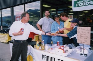 Man with a can of pop in hand, buying a hot dog at a table from four volunteers.