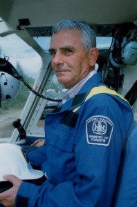 Man in blue coveralls seated with hard hat in hand, seated in a helicopter cabin.