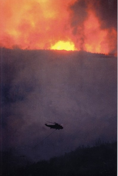 A helicopter hauling a water bucket approaches a blazing fire.
