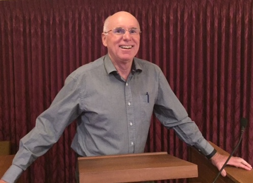 Man in a grey shirts smiles, standing at a lectern.