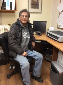 Man in a winter jacket sits at a computer desk, smiling.