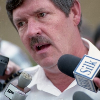 Man speaking into several microphones. Wears a white shirt.