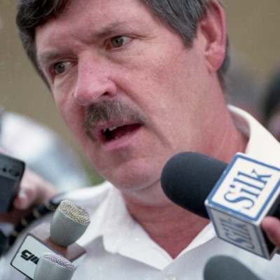 Man speaking into several microphones. Wears a white shirt.