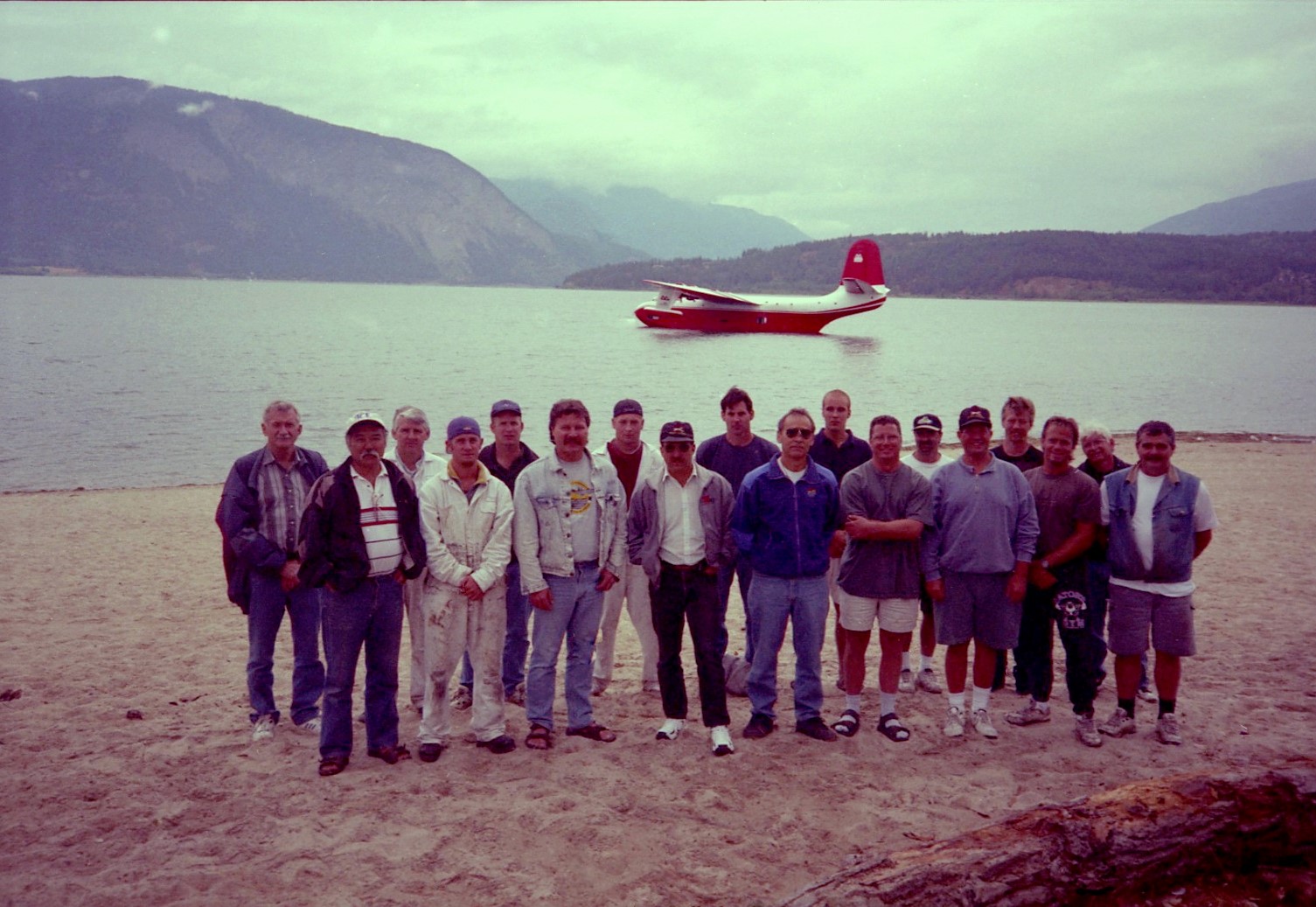 18 men stand on shore in front of a float aeroplane.