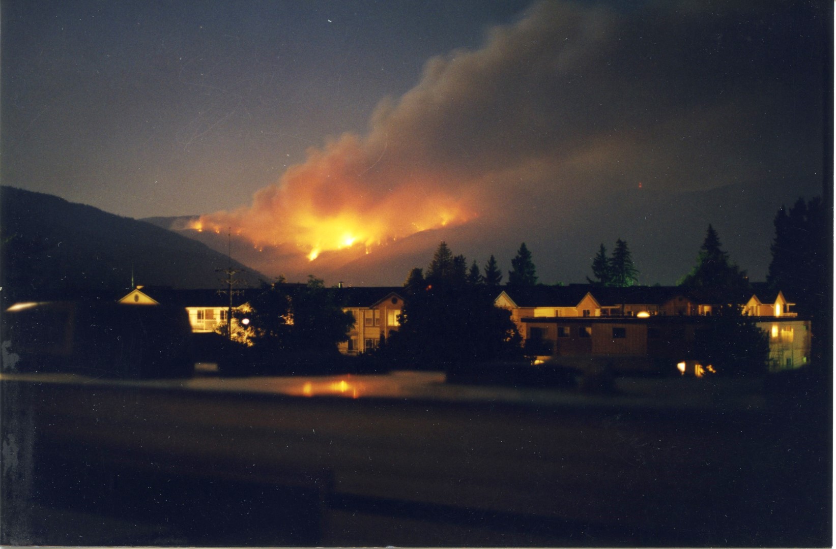 Fire burns bright red at night. Buildings lit in the foreground.