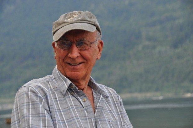 Man in a baseball cap smiles, lake and hillside in background.
